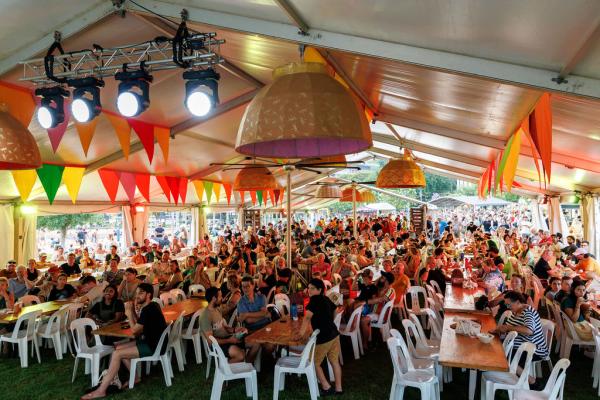 A big crowd of people are sitting at long tables and chairs underneath a large marquee. The ceiling of the marquee is decorated by colourful flag bunting, and large ornamental hanging lanterns. 