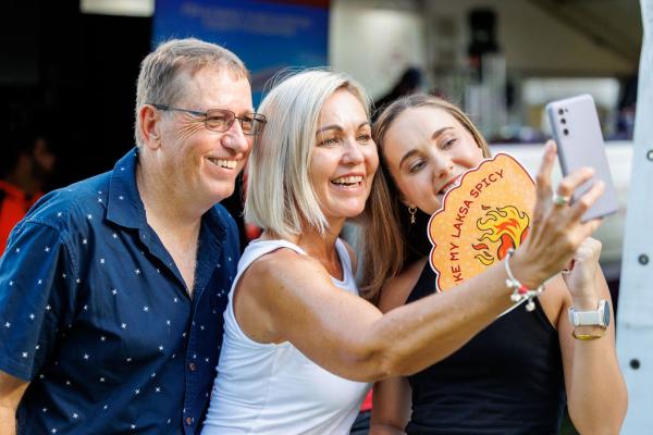 A group of 3 people posing for a selfie with a Laksa Festival handheld fan