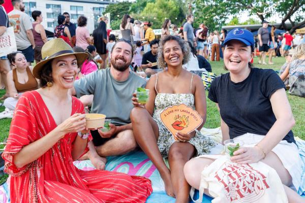 A group of 4 people sitting on a picnic rug, eating laksa and drinking cocktails