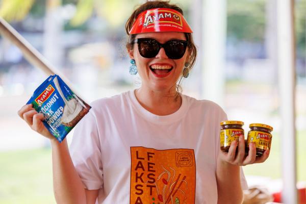 A lady holding up 2x jars of laksa paste in one hand, and a packet of coconut milk in the other hand, smiling